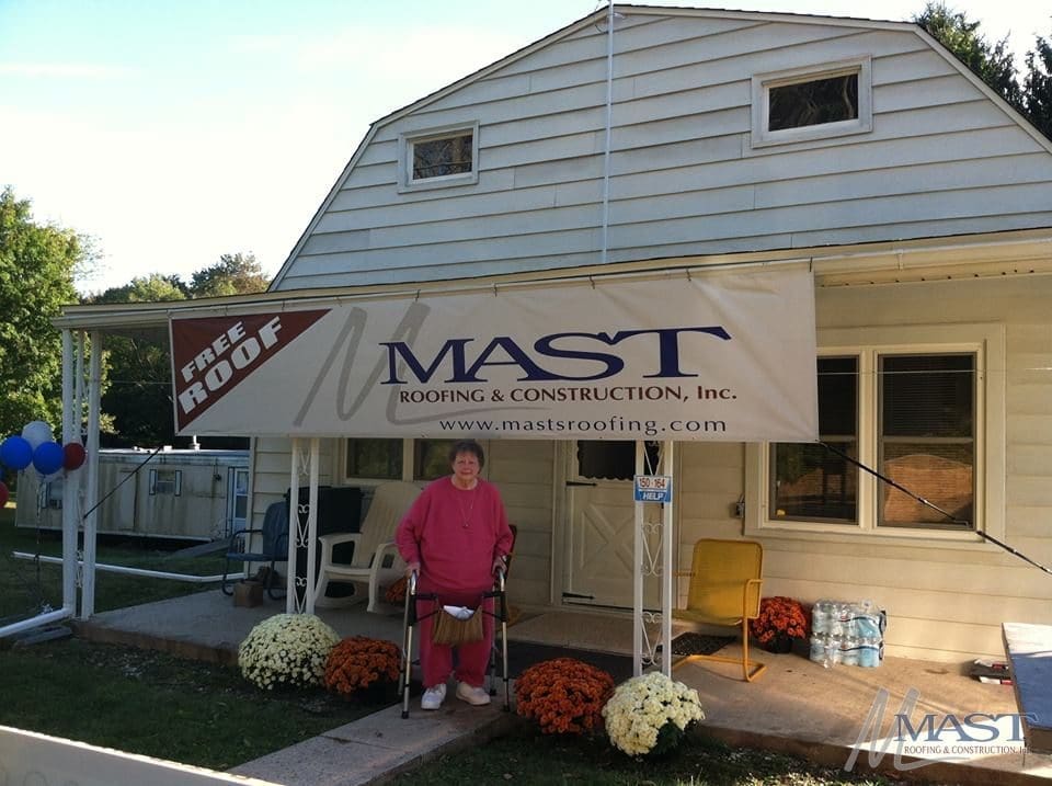 woman with a walker standing in front of a home with a new roof with a sign that says "Free Roof - Mast Roofing & Construction Inc"